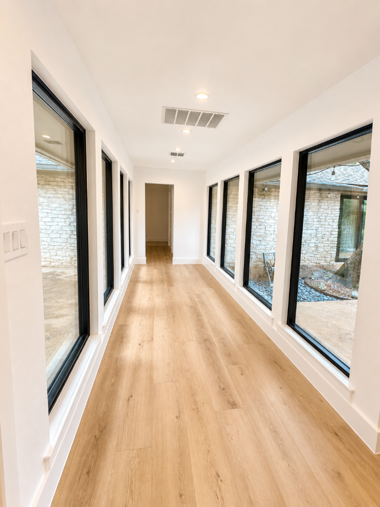 Symmetrical corridor with light oak plank flooring, black-framed floor-to-ceiling windows, and crisp white walls overlooking a stone courtyard
