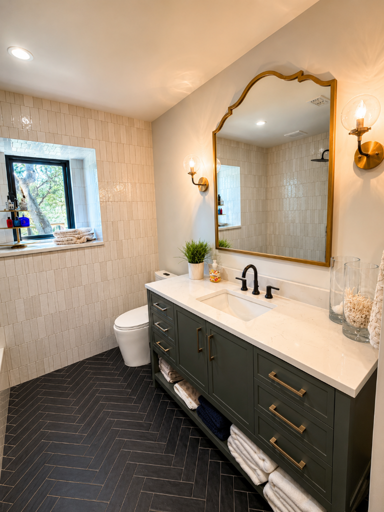 Forest green vanity with quartz countertop, arched brass mirror, and herringbone tile floors