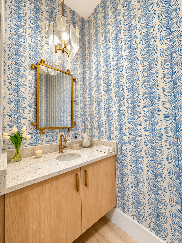 Blue fern wallpaper, oak vanity with marble countertop, and a gold geometric mirror with brass chandelier