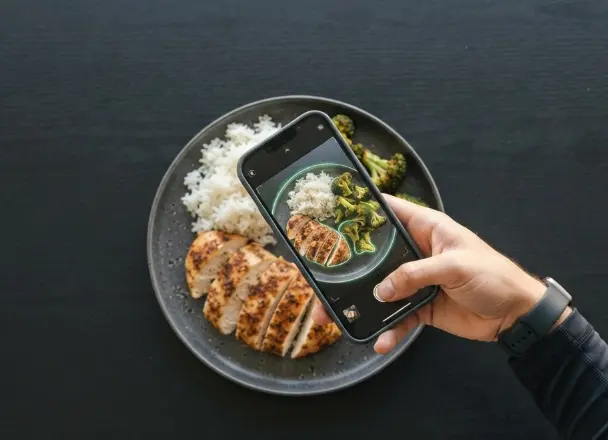 Top-down view of a hand holding a smartphone over a plate of sliced grilled chicken, white rice and broccoli while a food tracker app highlights each food