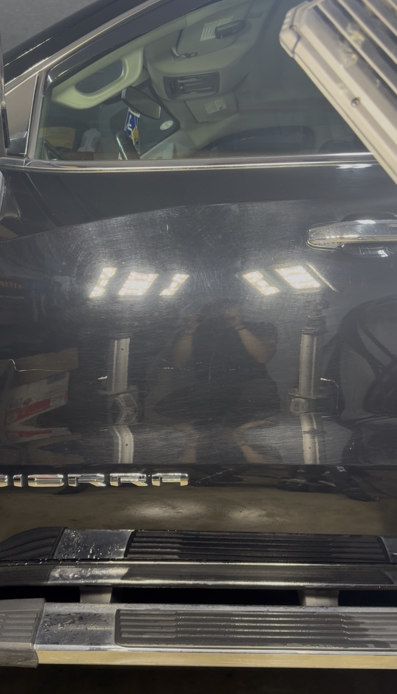 Close-up of a black GMC Sierra door panel covered in fine swirl marks and spider-webbing scratches under shop lighting