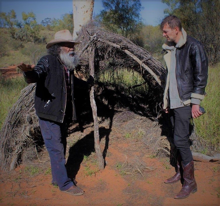 Elder explaining beside a traditional brush shelter
