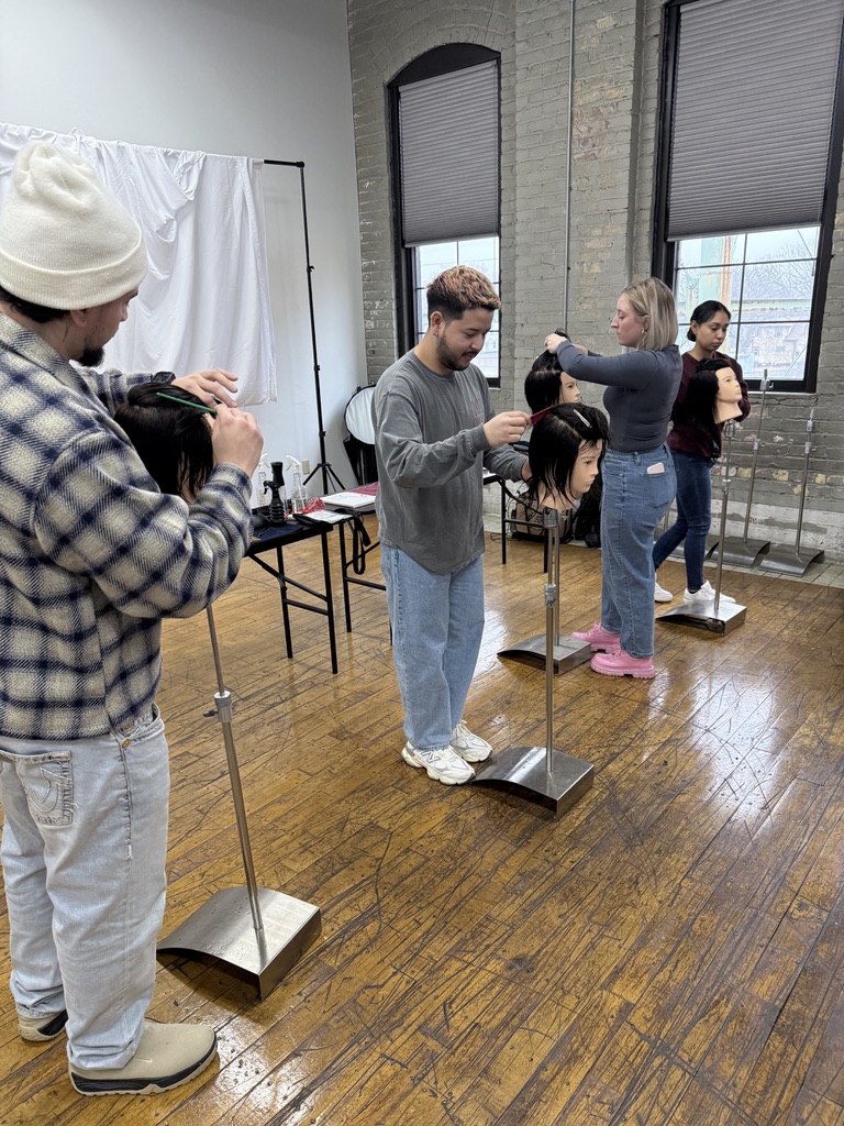 Stylists working on mannequin heads in a bright studio with large windows and exposed brick