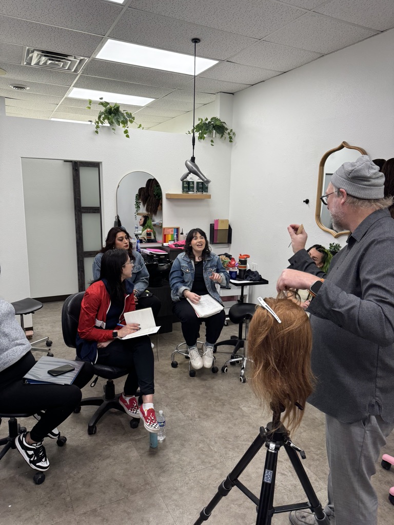 Instructor in a grey beanie teaching a group of seated stylists during a hands-on education session