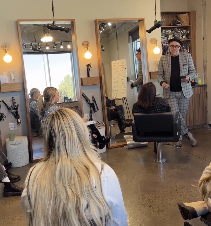 Instructor in a plaid jacket teaching a group of seated stylists in a salon classroom with large mirrors