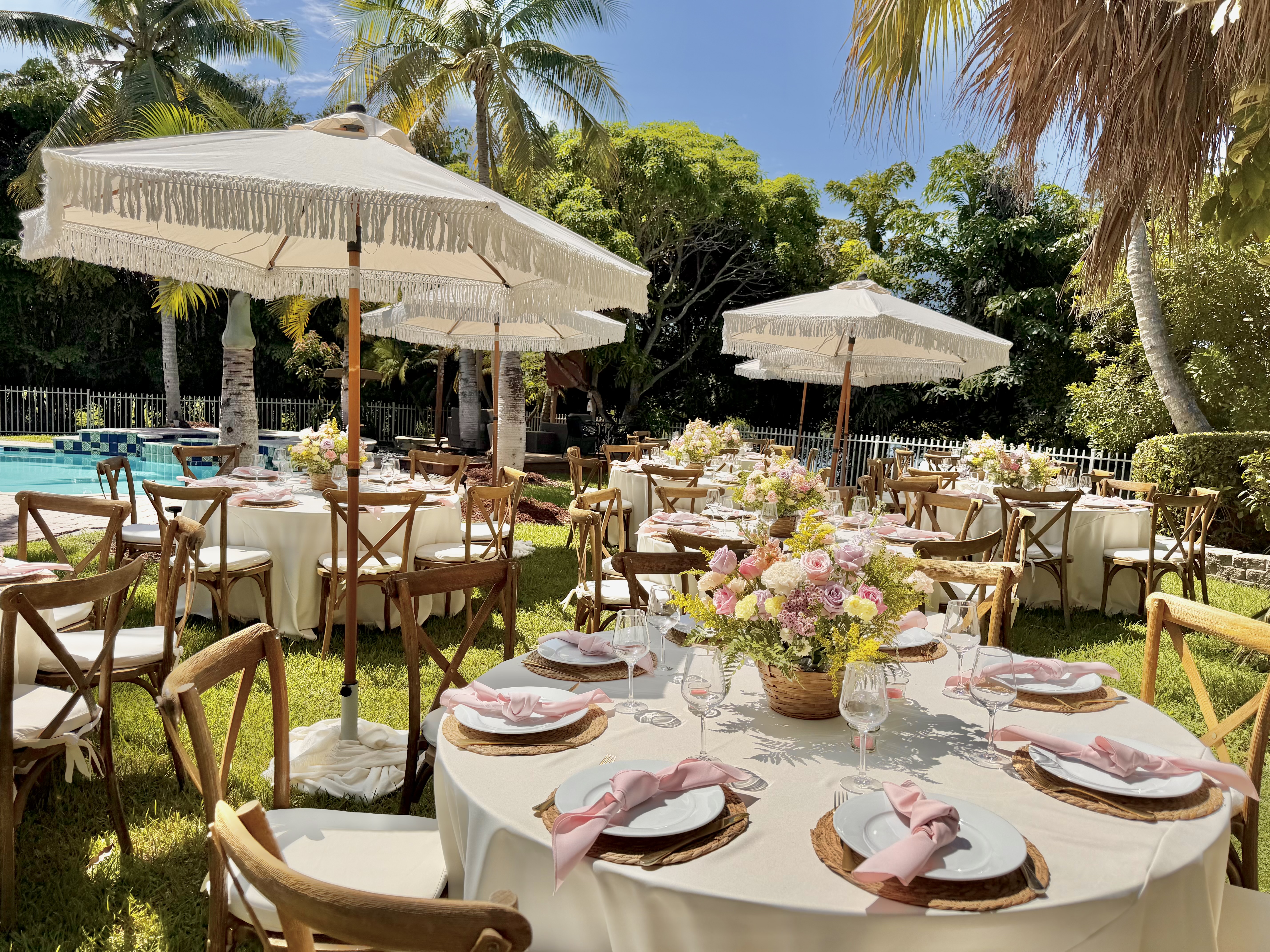 Outdoor event setup with round tables, white tablecloths, and umbrellas in a tropical backyard