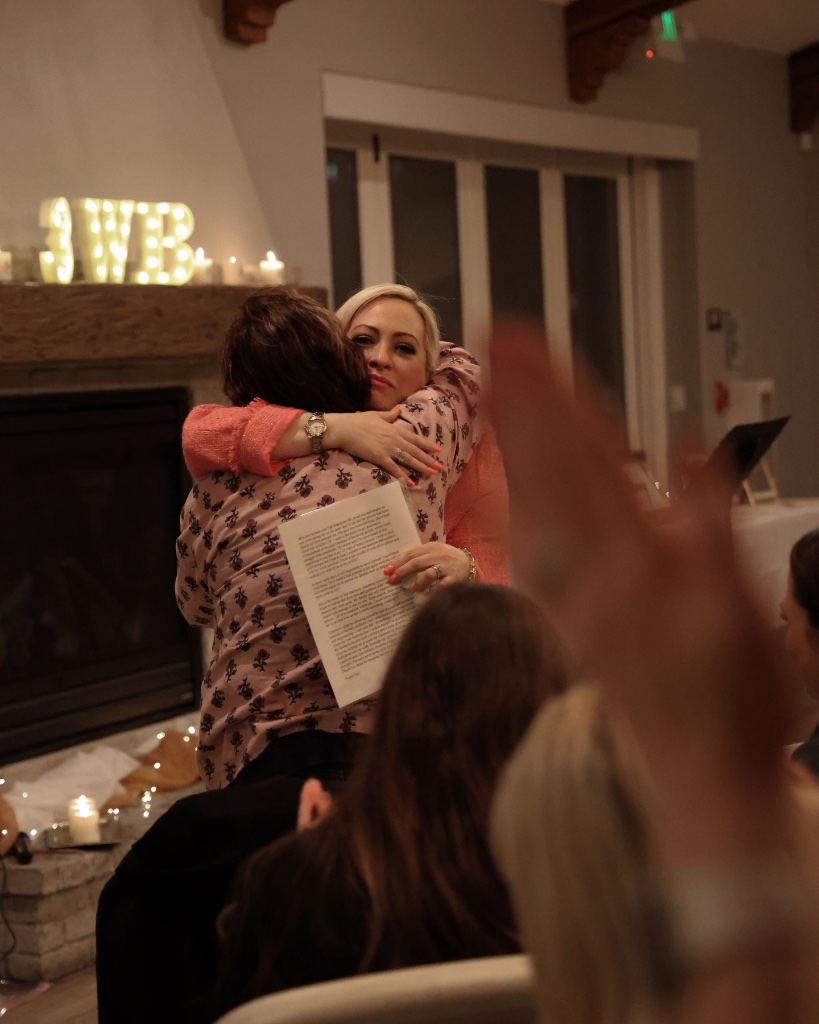 Two women sharing a warm embrace by a fireplace at a She Connects event