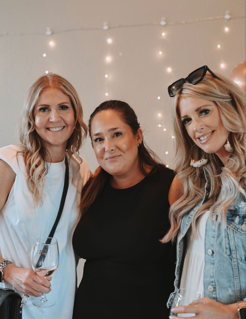 Three friends smiling with wine glasses at a She Connects event