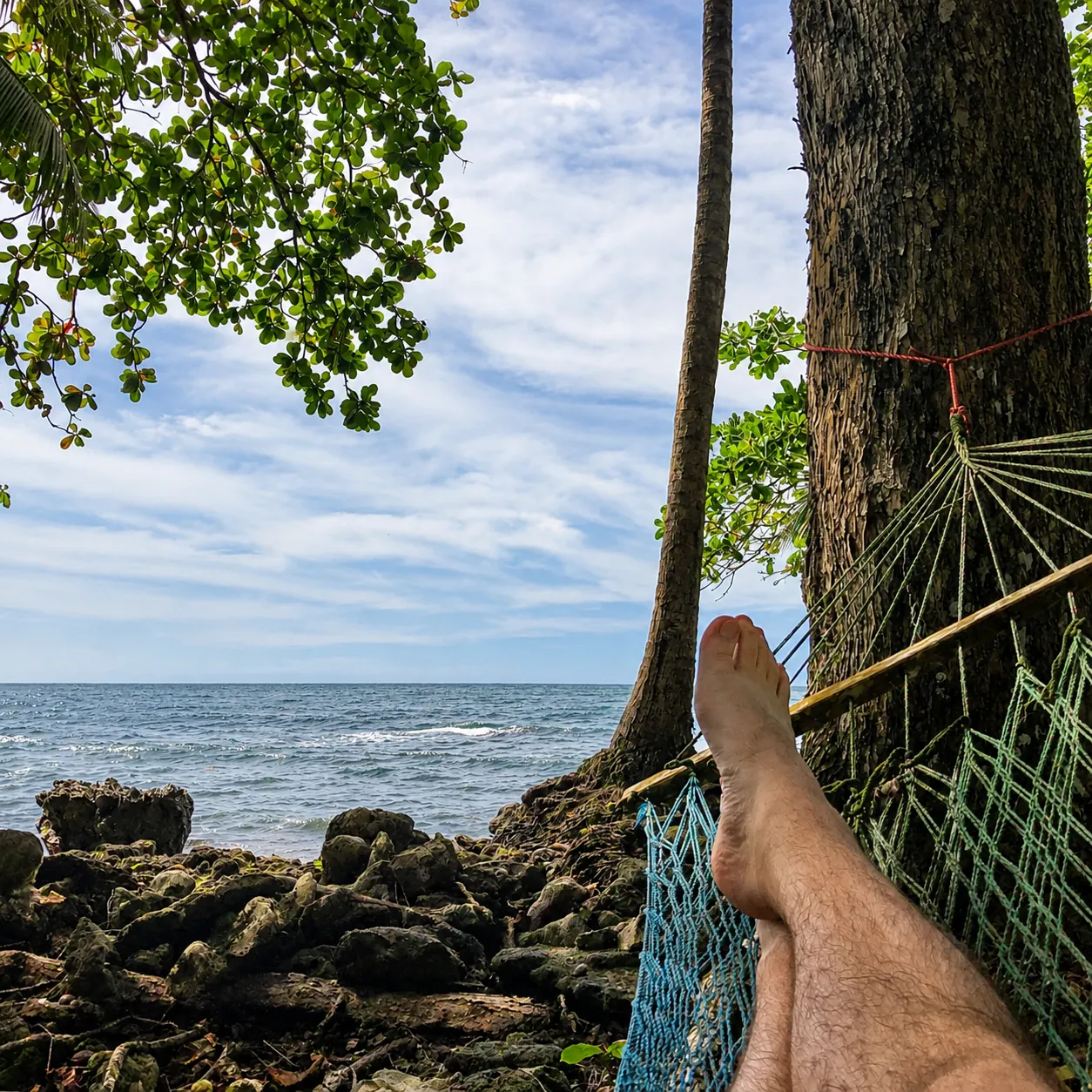 First-person view from a hammock beneath tropical trees, with crossed legs overlooking a rocky shoreline and the ocean.