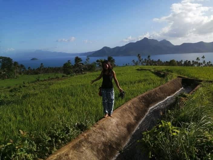 Guest standing beside a water channel with green rice fields and the sea beyond