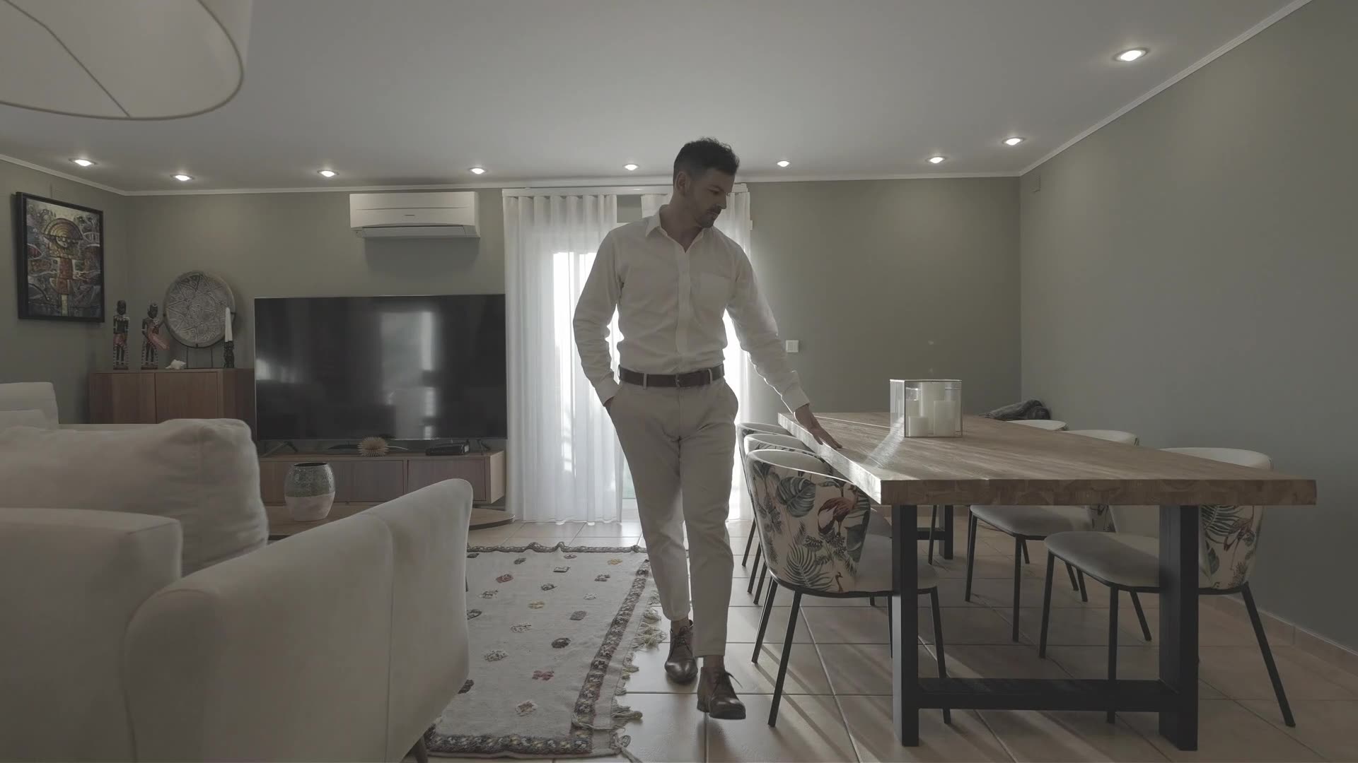 Benjamín Valdivia walking through a modern Lisbon apartment, resting his hand on a wooden dining table beside a sheer-curtained window