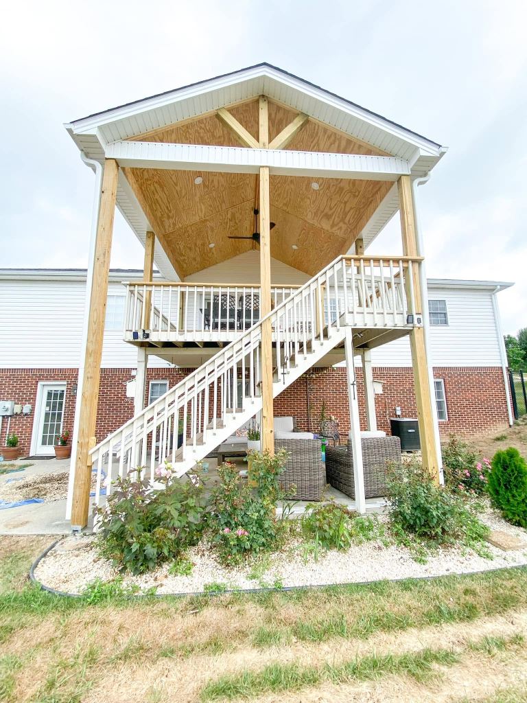 Custom-built two-story wood-framed covered deck and patio with natural lumber posts, white railing, and brick foundation