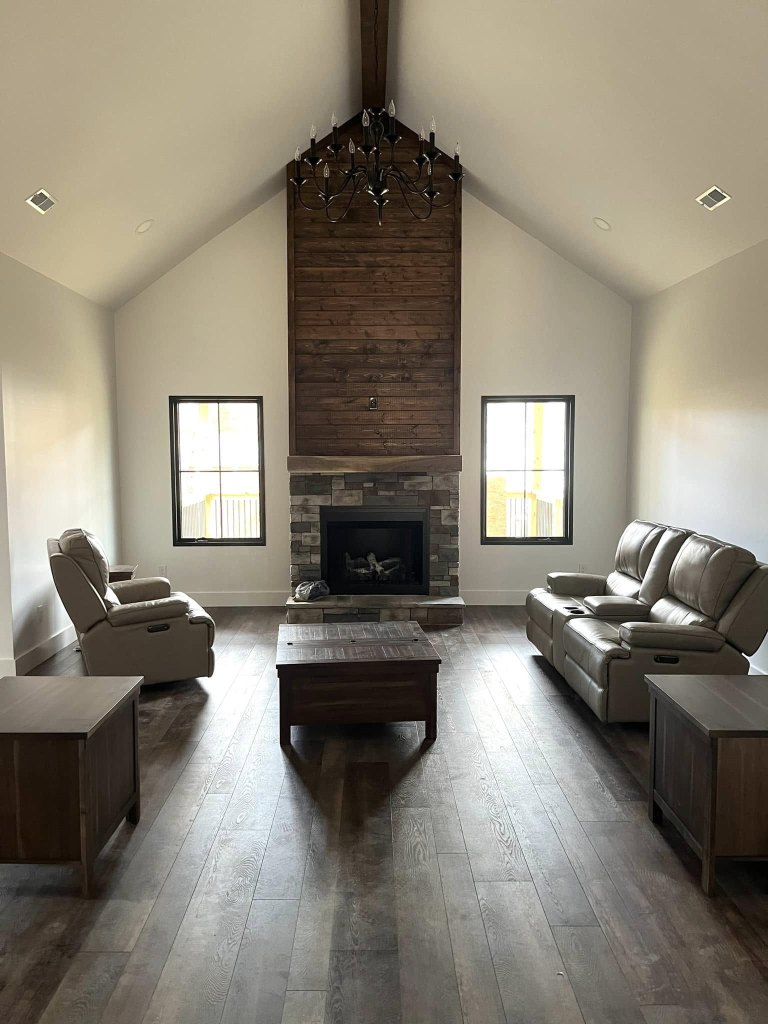 Modern rustic living room with vaulted ceiling, stacked-stone fireplace, dark wood accent wall, and leather seating