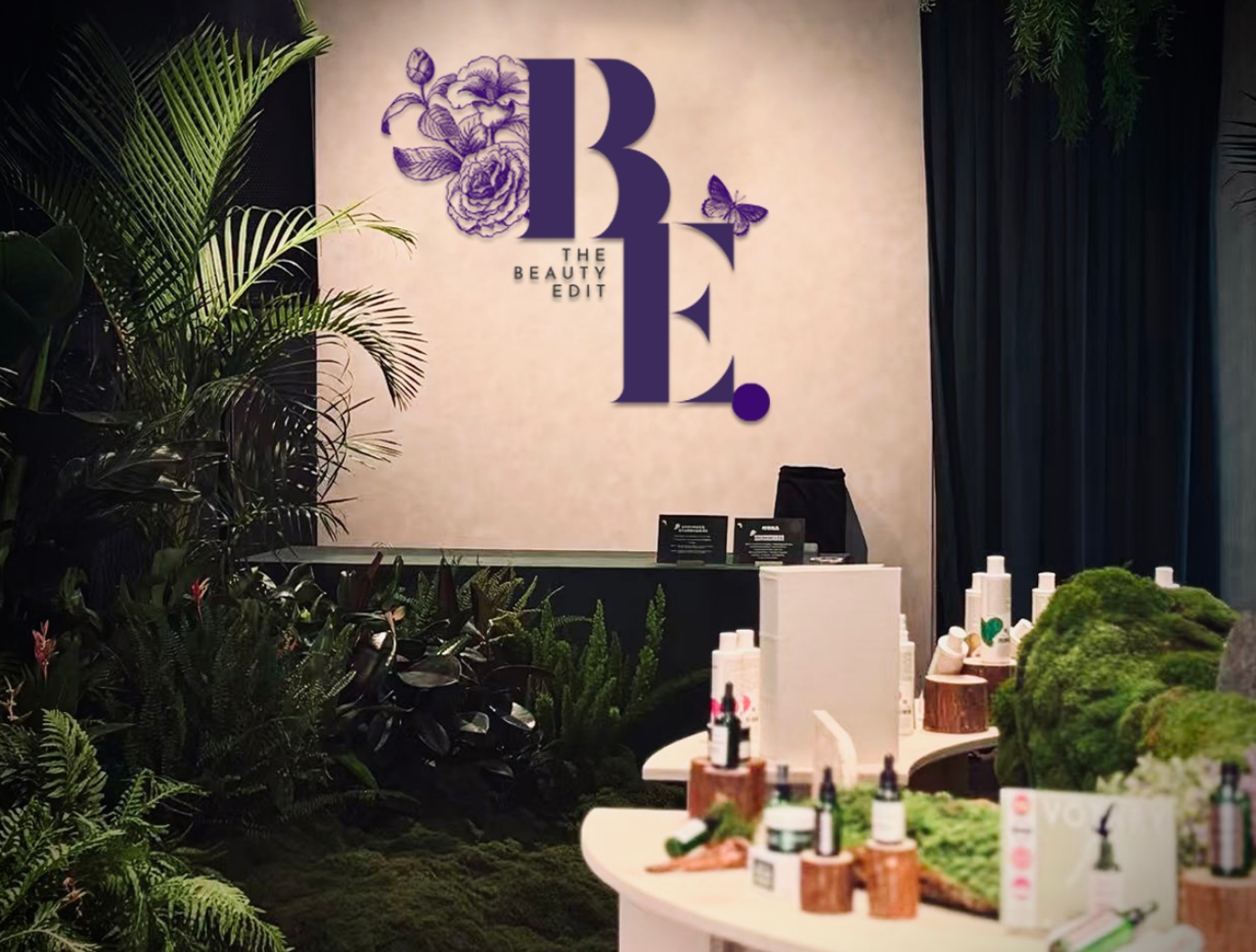 The Beauty Edit monogram wall above the reception counter, framed by ferns and moss