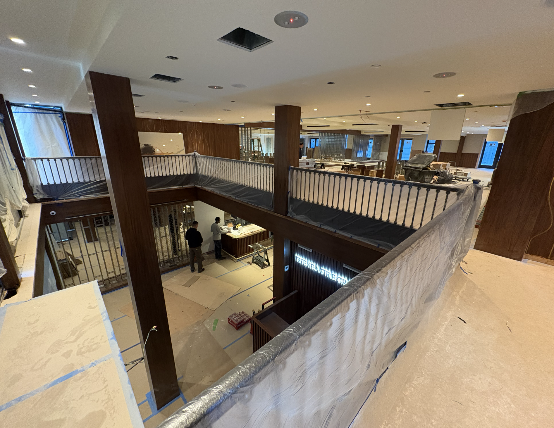 Two-level Seafire Grill restaurant interior under construction with walnut columns, a wrapped mezzanine railing and an illuminated Seafire sign below at the Seafire Grill 49th Street location in NYC