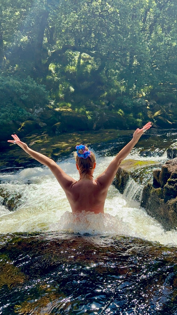 A woman immersed in a wild forest river, arms raised in celebration