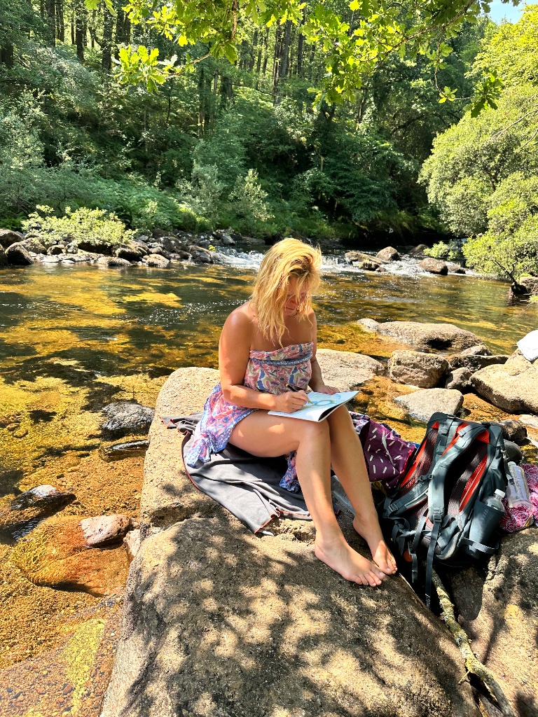 Vittoria Immortelle painting in the luminous living field, seated on a rock in a sunlit river