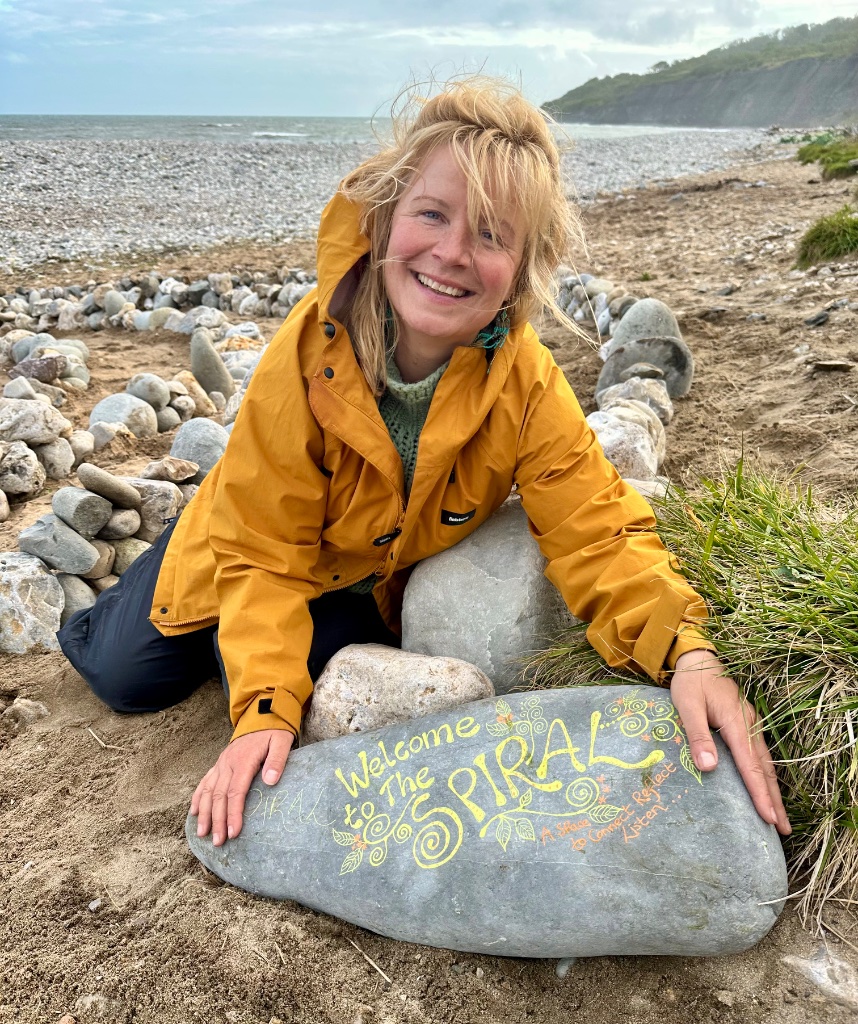 Vittoria Immortelle kneeling beside a hand-painted spiral stone on a wild beach