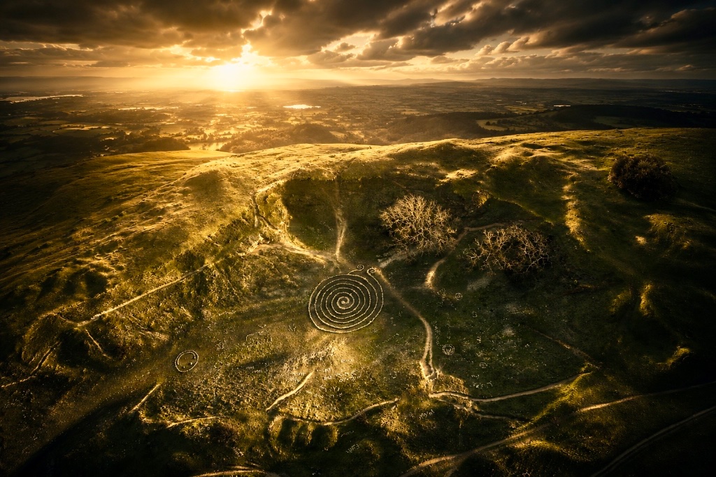Sacred stone spiral land art on a golden-hour hillside, by land artist Vittoria Immortelle