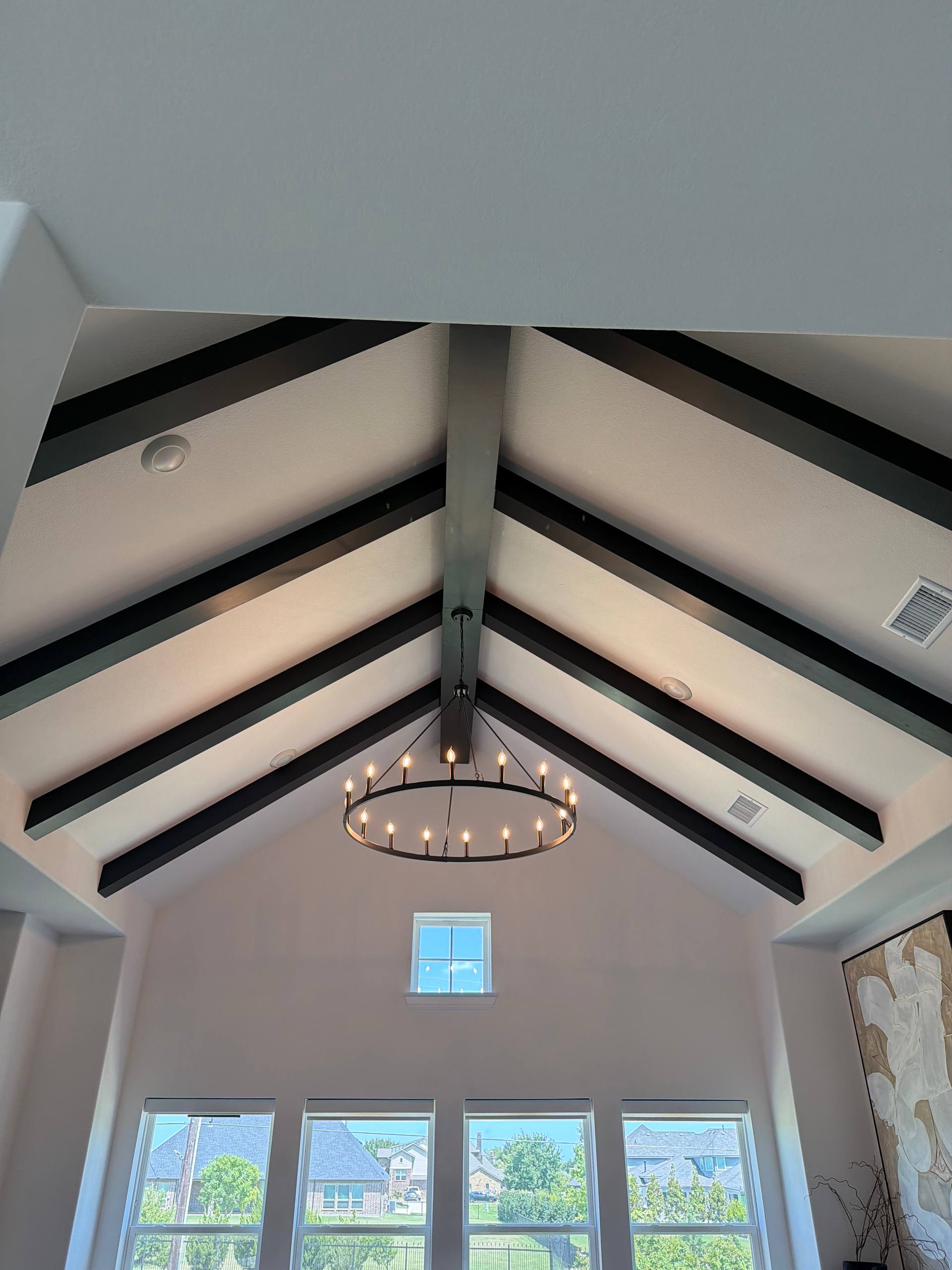Vaulted ceiling with dark charcoal exposed wood beams meeting at a central ridge beam, a circular wagon-wheel chandelier, and a row of large windows in a North Texas home