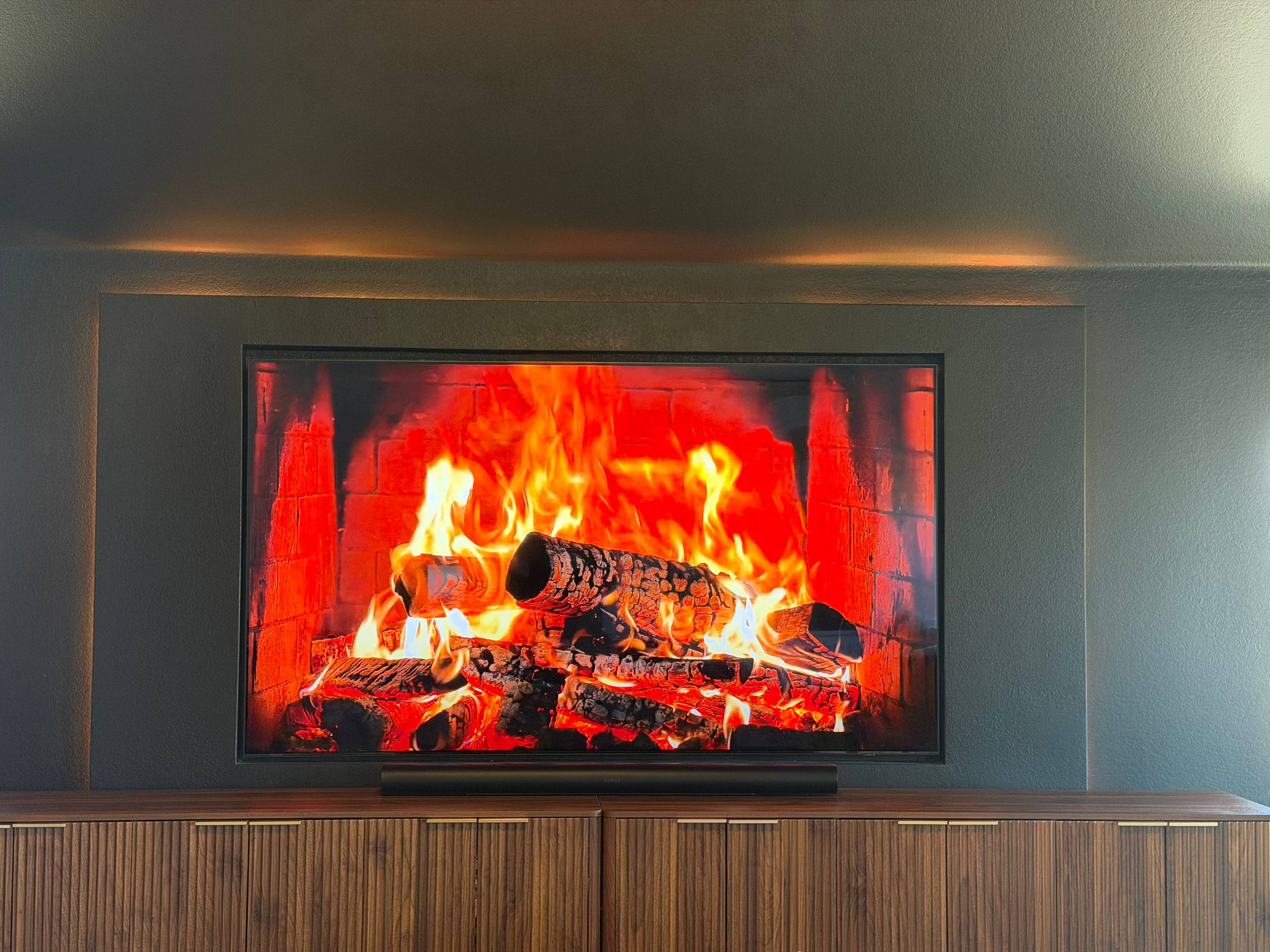 Wall-mounted TV above a reeded white oak console with brass handles on a charcoal backlit wall