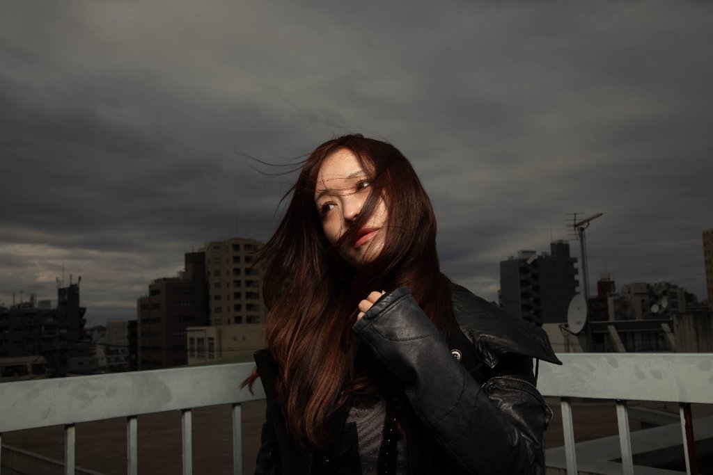 A high-resolution portrait of a woman with windblown chestnut-brown hair on an urban rooftop with dramatic overcast grey sky and cityscape