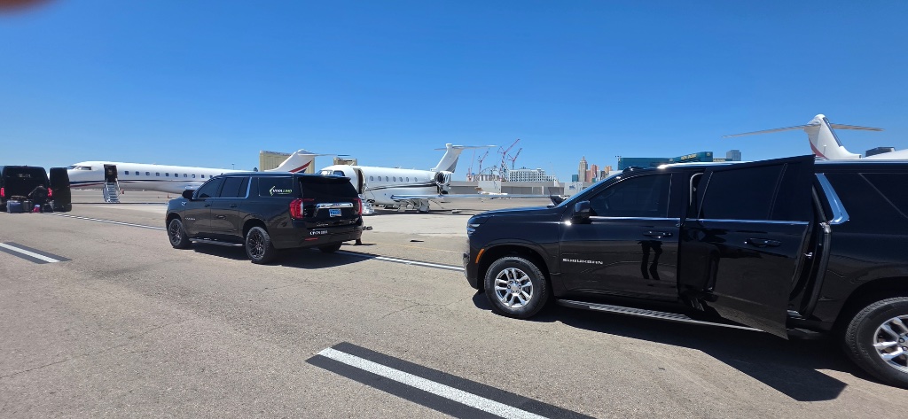 Two black Suburban SUVs staged on an airport tarmac beside private jets