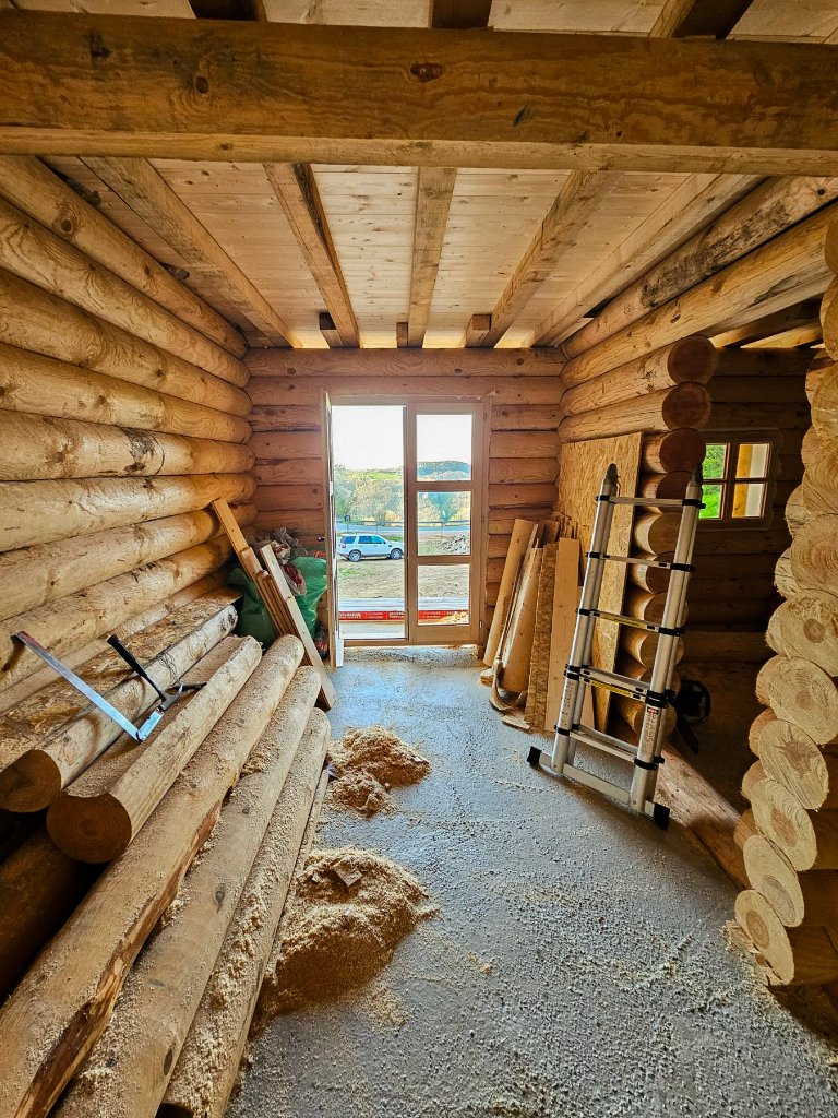 Enterijer brvnare u izgradnji — betonski pod, složena brvna i otvorena vrata