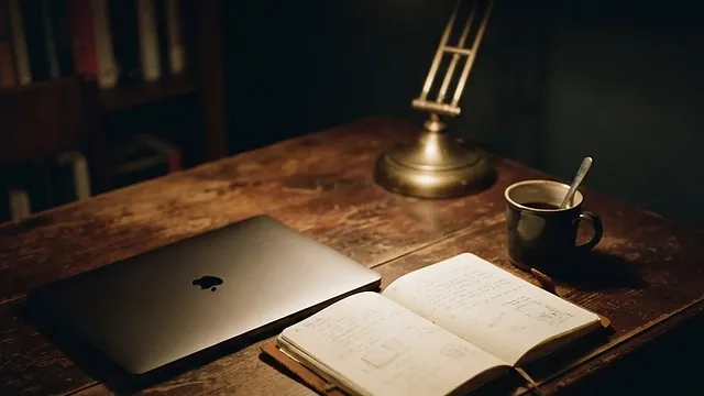 A desk at night under a single lamp, a closed laptop beside an open notebook.