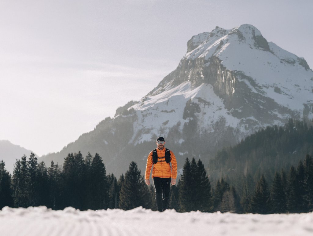 Man in an orange outdoor jacket walking through a snowy mountain landscape