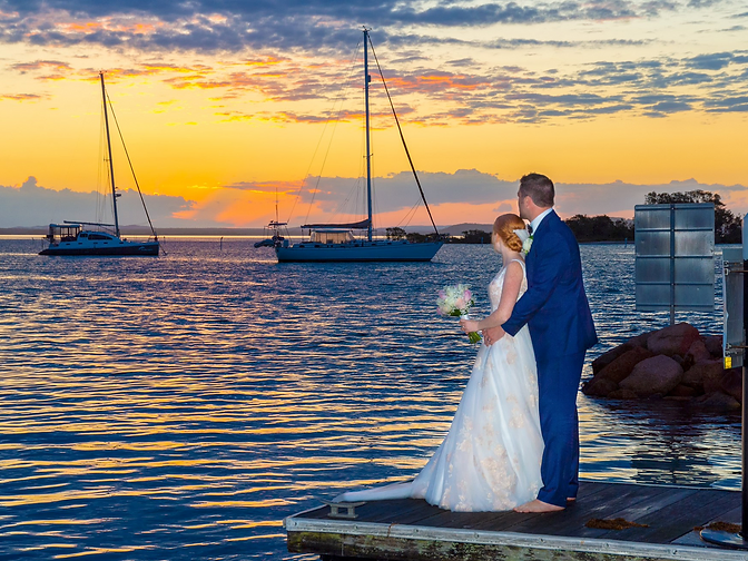 A couple standing on a wooden jetty at sunset looking out over the calm Port Stephens bay, with sailboats silhouetted against the golden horizon