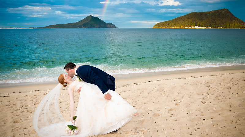 A coastal wedding ceremony on the Port Stephens shoreline at golden hour, the couple framed by the bay and soft warm light