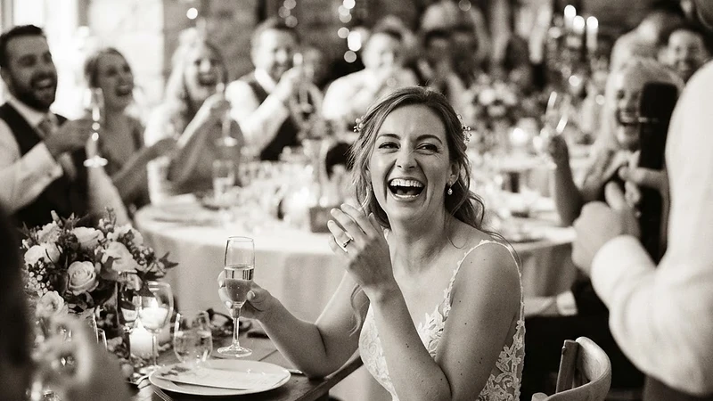 A candid documentary wedding moment — a bride laughing naturally during a reception speech, warm natural light, unposed authentic emotion