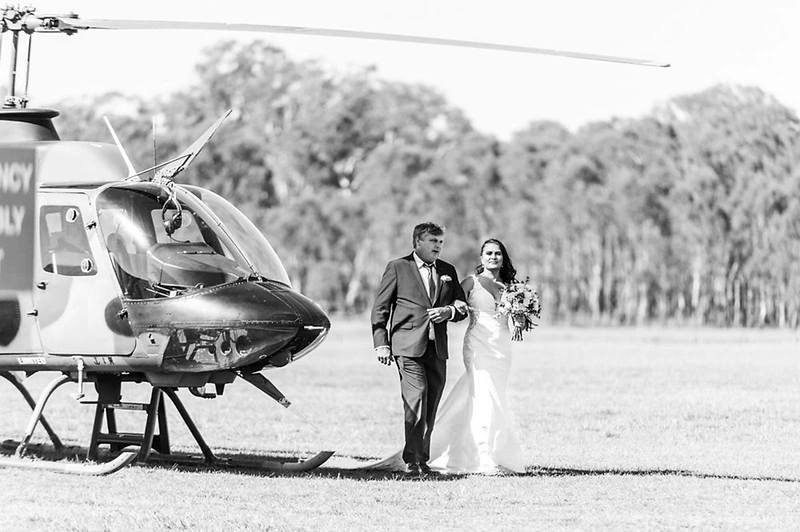 Rachel and Greg walking away from a helicopter across the grass airfield at Luskintyre in the Hunter Valley — a real Hunter Valley wedding photographed by Newcastle wedding photographer John Adams