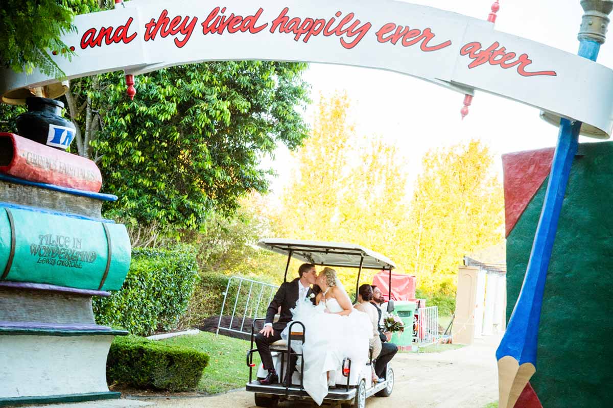 Stacey and Michael kissing in a golf cart framed by a fairytale storybook archway of stacked oversized books and a giant pencil at their Hunter Valley Gardens wedding — a real Hunter Valley wedding photographed by Newcastle wedding photographer John Adams