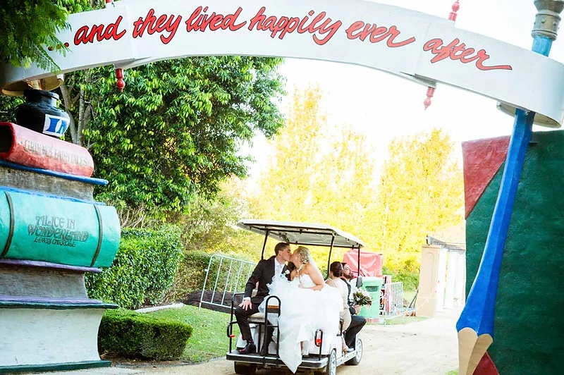 Stacey and Michael kissing in a golf cart framed by a fairytale storybook archway of stacked oversized books and a giant pencil at their Hunter Valley Gardens wedding — a real Hunter Valley wedding photographed by Newcastle wedding photographer John Adams