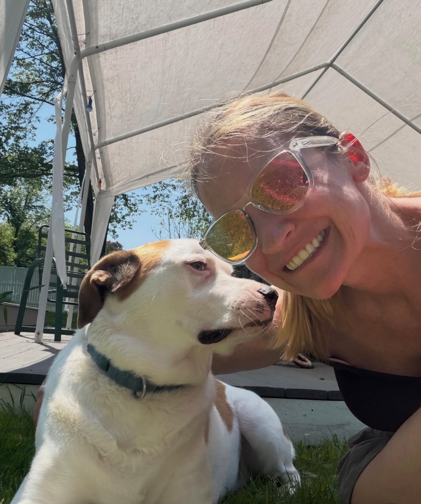 Casey smiling cheek-to-cheek with a white and brown dog outdoors on a sunny day