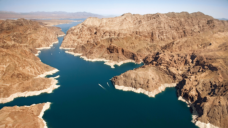 A deep-blue reservoir winding through arid desert mountains, with a visible shoreline mineral ring.