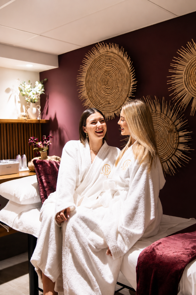 Two spa guests laughing together in robes against an oxblood wall