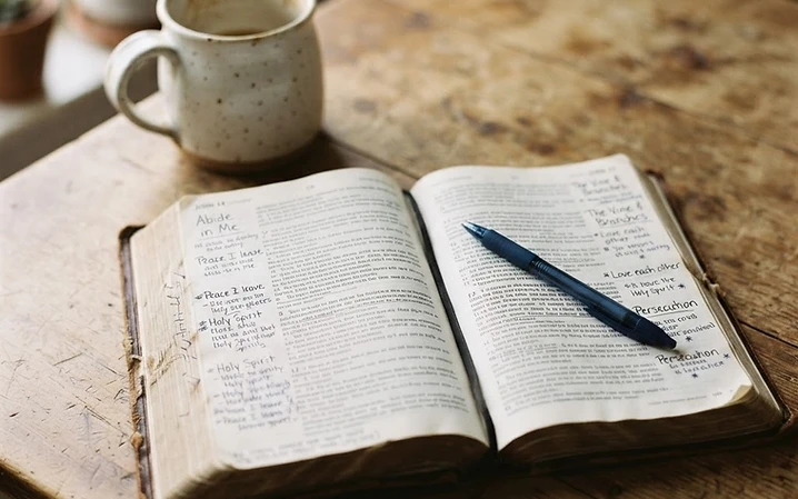 An open Bible with handwritten notes in the margins on a wooden table in soft morning light