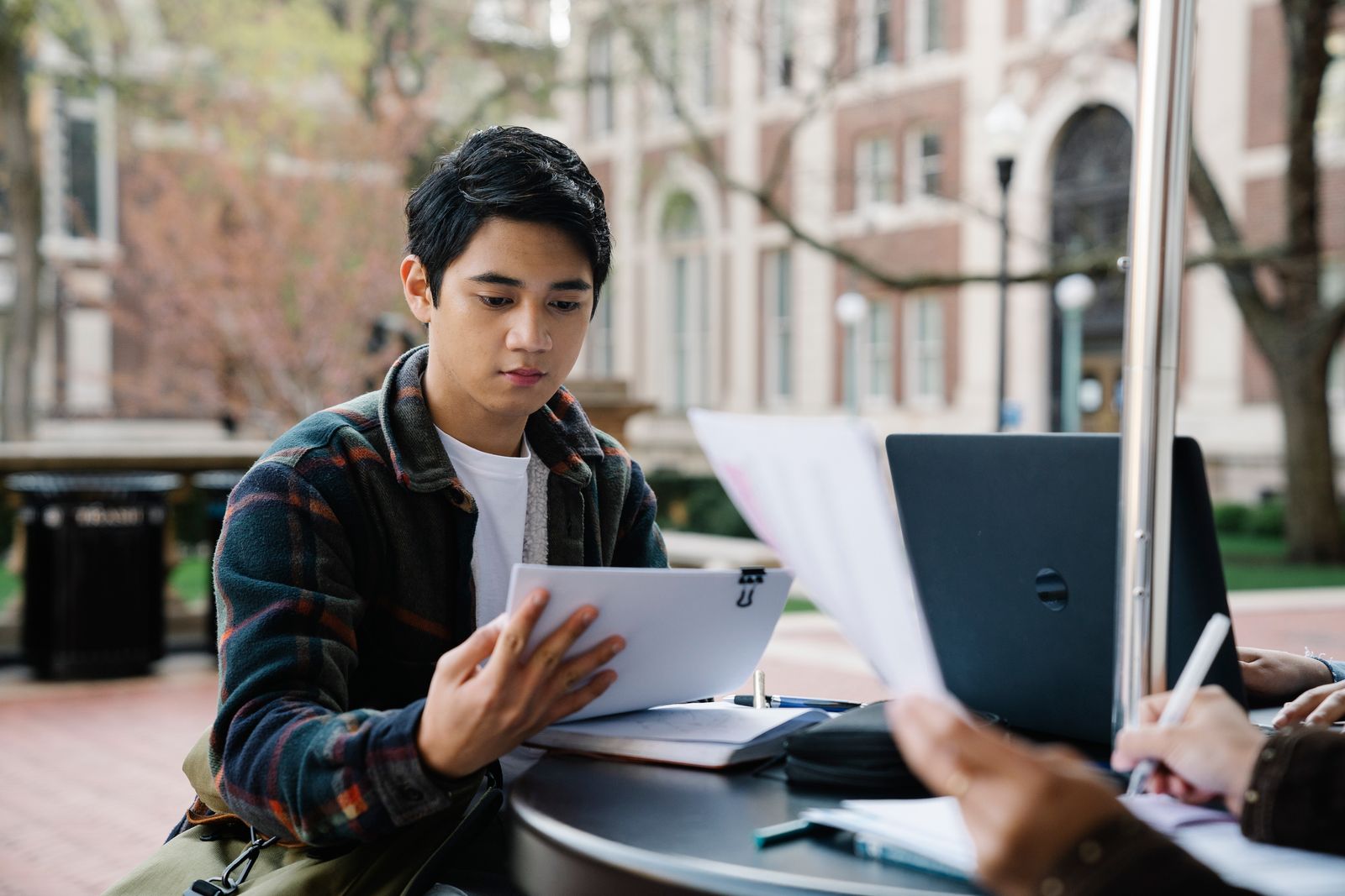 A college student studying papers at an outdoor table on a campus