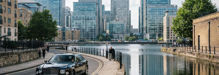 Black executive saloon approaching Canary Wharf with glass skyscrapers reflected in dock water in bright natural daylight