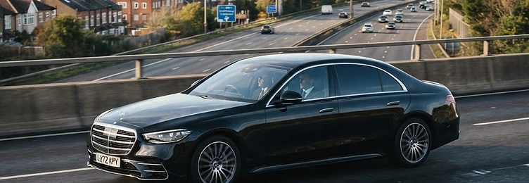 Black executive saloon on a motorway with suburban London rooftops in the distance in bright natural daylight