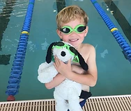 A young boy wearing green swim goggles hugs a stuffed dog toy wearing matching goggles at the edge of a swimming pool