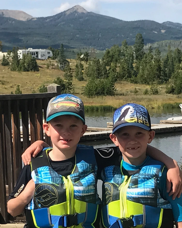 Two children wearing brightly colored life jackets beside a mountain lake