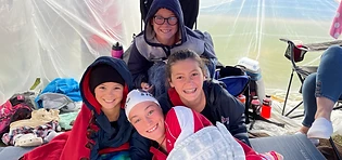 Four young swimmers huddled together in blankets and hoodies under a clear tent at a swim meet