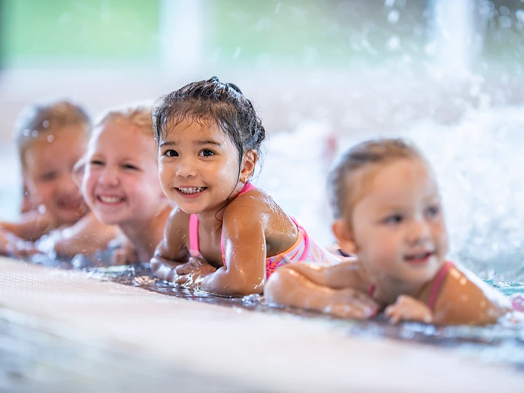 Four smiling children resting their arms on the deck of a bright indoor pool during a swim session