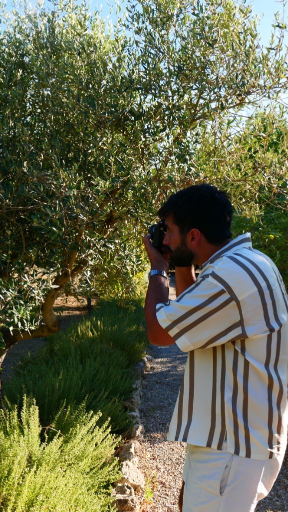 Jake in profile looking through a camera beside an olive tree