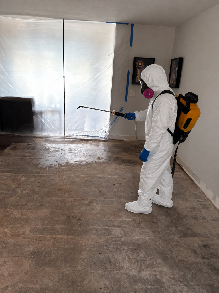 Technician in full PPE applying antimicrobial treatment inside a contained, plastic-sealed room
