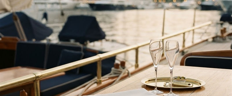 A teak yacht deck at golden hour, linen napkin and crystal glasses, harbour beyond.